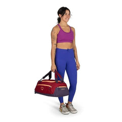 The image shows a person standing against a plain background, dressed in athletic attire. They are wearing a purple sports bra and blue leggings, paired with gray sneakers that have red accents. The person is holding a red and purple gym bag in their righ