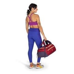The image shows a woman standing and facing away from the camera, dressed in athletic wear. She is wearing a magenta sports bra with cross-back straps, and vivid blue high-waisted leggings that have pocket details on the sides. The woman has several tatto