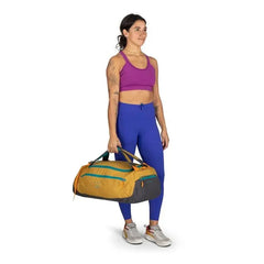 The image shows a person standing and holding a large, stylish duffle bag. The individual is wearing athletic attire, including a vibrant purple sports bra and blue leggings, paired with light-colored sneakers. The duffle bag is prominently designed with