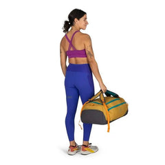 The image features a woman standing and facing away from the camera. She is wearing a vibrant sports outfit, which includes a purple sports bra and blue leggings with a stripe detail. Her hair is tied back in a ponytail, and she has visible tattoos on her