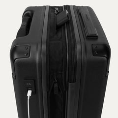 Close-up of the external charging ports on the Travelpro Platinum Elite Compact Carry-On Hardside Spinner in Shadow Black, showing a white cable plugged into the USB port and a power bank tucked into the dedicated battery sleeve within the expansion gusset.
