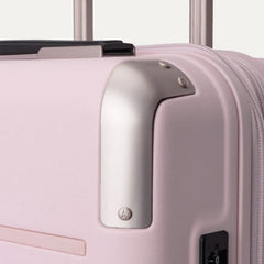 Close-up view of the light pink Travelpro Platinum Elite Next Gen Powder Pink Suitcase, highlighting the metallic corner guard, top carry handle, zipper, and telescoping handle against an off-white background.