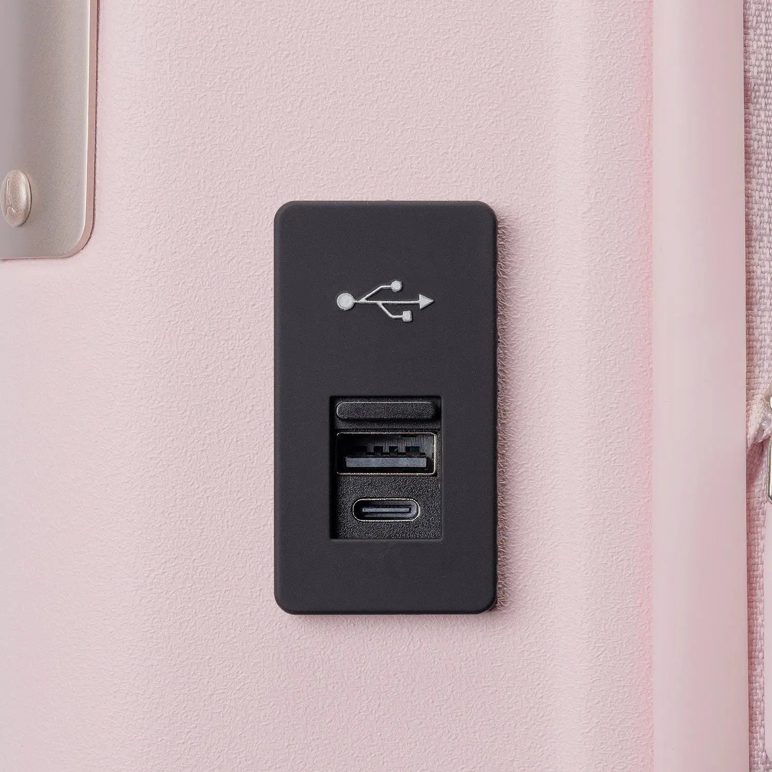 A black rectangular USB port cover plate with integrated charging ports, mounted on a light pink textured surface of the Travelpro Platinum Elite Next Gen Powder Pink Suitcase.