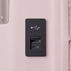 A black rectangular USB port cover plate with integrated charging ports, mounted on a light pink textured surface of the Travelpro Platinum Elite Next Gen Powder Pink Suitcase.