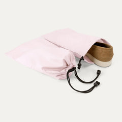 A Powder pink shoe bag with black drawstrings, shown with a tan canvas shoe on the end, on a clean off-white background.