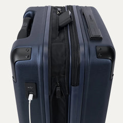 Close-up of the external charging ports on the Travelpro Platinum Elite Carry-On Hardside Spinner in Aviator Navy, showing a white cable plugged into the USB-A port and a power bank tucked into the dedicated battery sleeve within the expansion gusset.