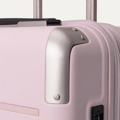 Close-up view of the light pink Travelpro Platinum Elite Next Gen Powder Pink Suitcase, highlighting the metallic corner guard, top carry handle, zipper, and telescoping handle against an off-white background.