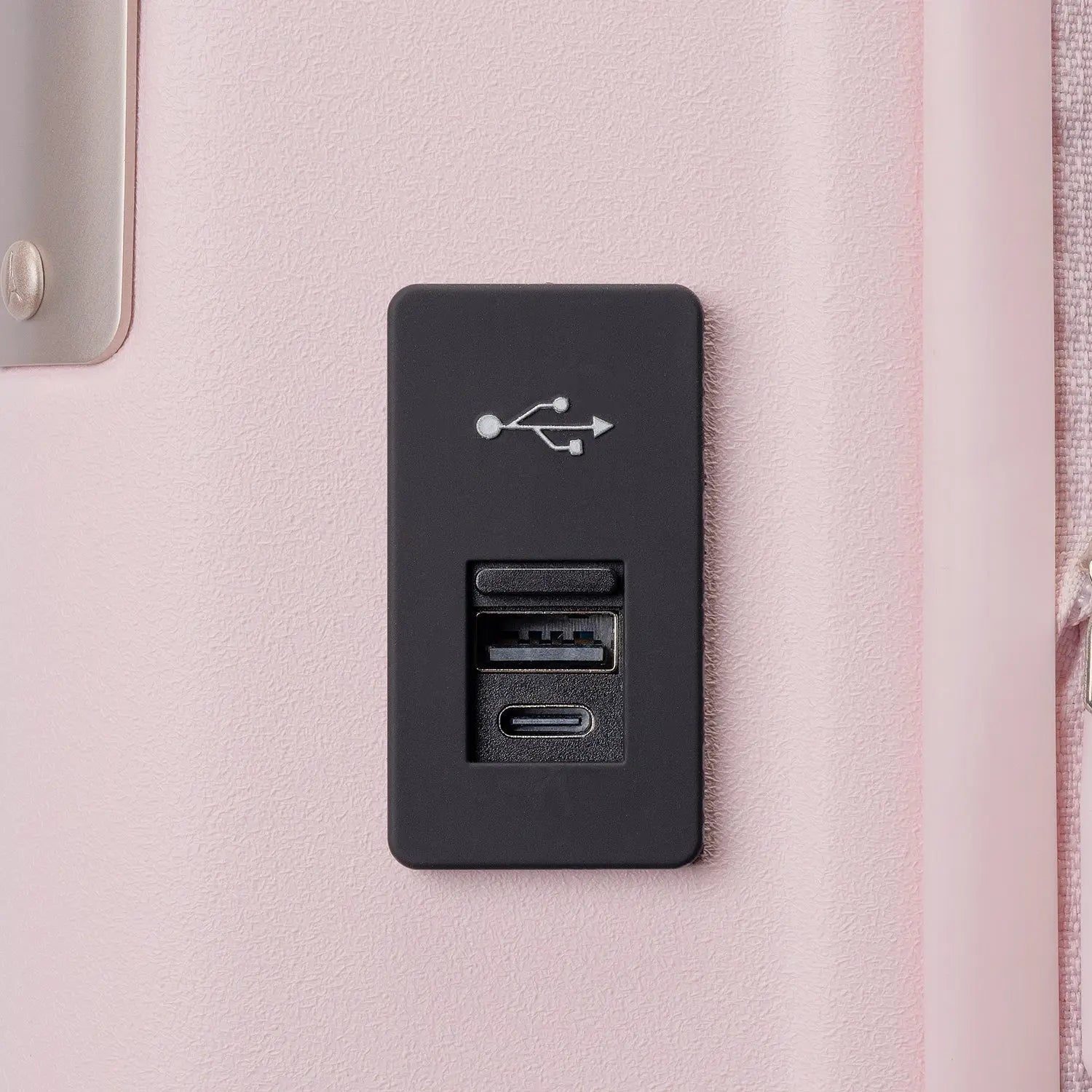 Close-up view of the black rectangular USB charging port panel mounted on the light pink Travelpro Platinum Elite Next Gen Powder Pink Suitcase, featuring a USB symbol and charging ports against an off-white background.