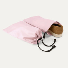 A Powder pink drawstring shoe bag with black cords, shown with a tan suede shoe partially tucked inside, on a clean off-white background.
