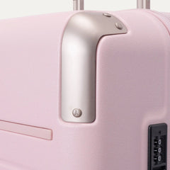Close-up of the Travelpro Platinum Elite Medium Check-In Hardside Spinner in Powder Pink, featuring protective polished aluminum corner armor, an integrated TSA-approved combination lock, and smooth side zipper trim against a clean off-white background.